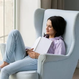 Mulher sentada na poltrona com um dos pés em cima do assento e com um livro sobre o corpo.